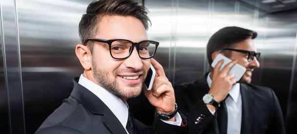panoramic shot of smiling businessman in suit talking on smartphone in elevator - Photo, Image
