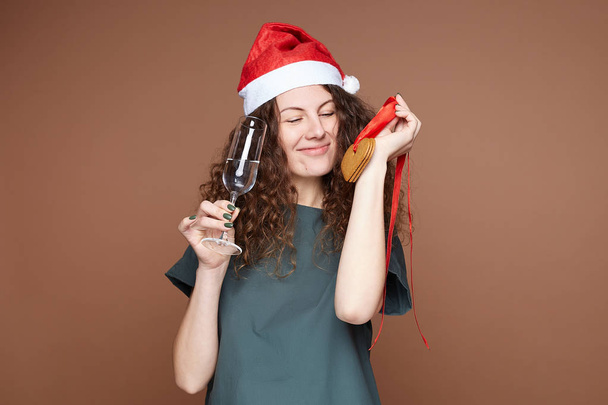 Attractive  young woman in Santa hat with glass of water holding handmade ginger cookies for Christmas tree decoration, can not wait to try freshly baked sweets, has curly hairstyle, not keep to diet. - Фото, изображение