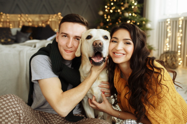 happy couple is sitting near the bed with a dog, christmas. new year at home. family and date - Φωτογραφία, εικόνα