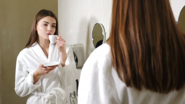 Giovane donna attraente in accappatoio bianco bere caffè davanti allo specchio del bagno. I filmati della vista posteriore. Bella donna guardando il suo riflesso nello specchio del bagno e sorridendo mentre si gode la mattina
 - Filmati, video