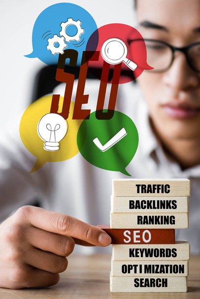 selective focus of asian seo manager holding wooden rectangle with lettering seo and sitting near seo illustration - Photo, Image
