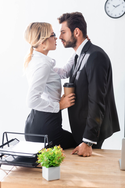 Πλευρική άποψη της σέξι επιχειρηματίας κρατώντας καφέ και αγκαλιάζει τον επιχειρηματία στο γραφείο - Φωτογραφία, εικόνα