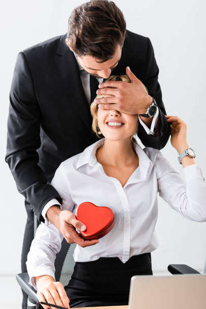 Businessman holding gift box and covering eyes to smiling colleague - 写真・画像
