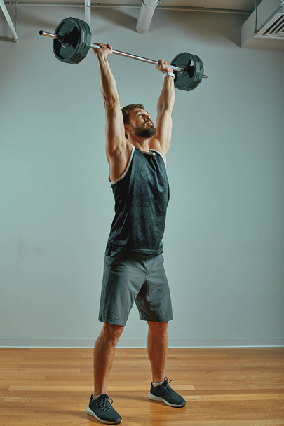 Porträt eines superfitten, muskulösen jungen Mannes, der im Fitnessstudio mit Langhantel auf grauem Hintergrund trainiert, Kopierraum. - Foto, Bild