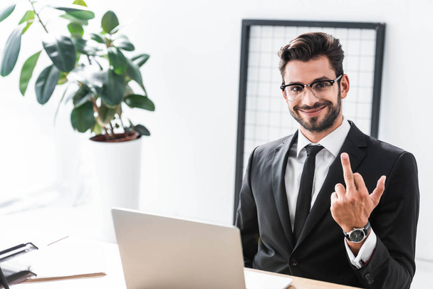 Smiling businessman showing middle finger gesture at office table - Foto, afbeelding
