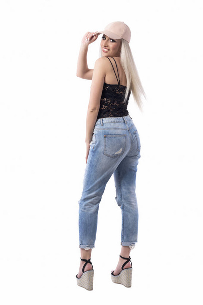 Back view of beautiful playful blond hair in jeans and cap holding visor turning and smiling at camera. Full body isolated on white background. - Photo, Image