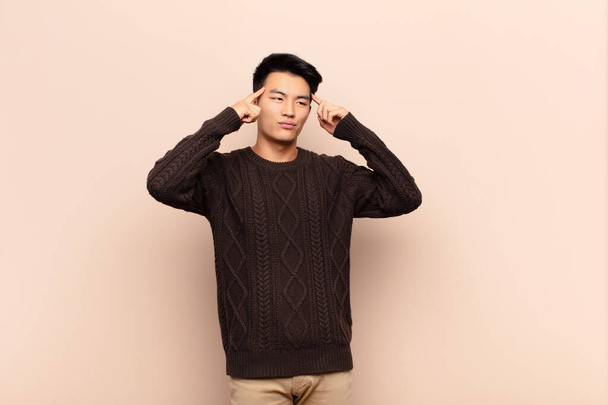 young chinese man feeling confused or doubting, concentrating on an idea, thinking hard, looking to copy space on side against flat color wall - Foto, afbeelding