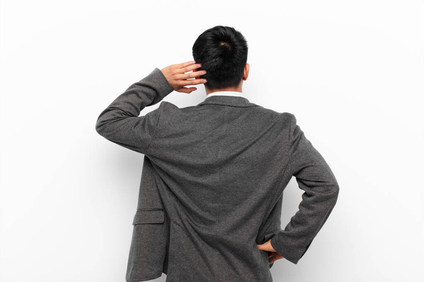 young chinese man thinking or doubting, scratching head, feeling puzzled and confused, back or rear view against flat color wall - Photo, image