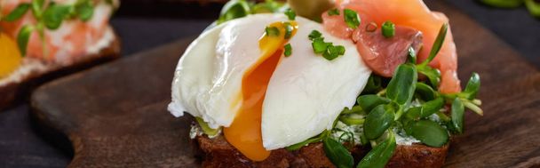 panoramic shot of danish smorrebrod sandwich with poached egg near salmon on wooden cutting board  - Fotografie, Obrázek