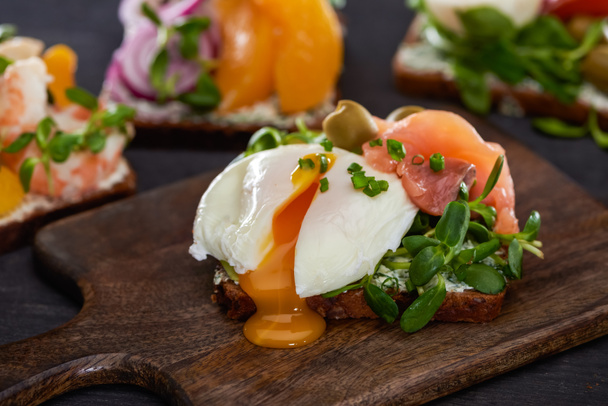κοντινό πλάνο του Danish Smorrebrod σάντουιτς με αυγό ποσέ κοντά σε φρέσκο σολομό σε ξύλινη επιφάνεια κοπής  - Φωτογραφία, εικόνα