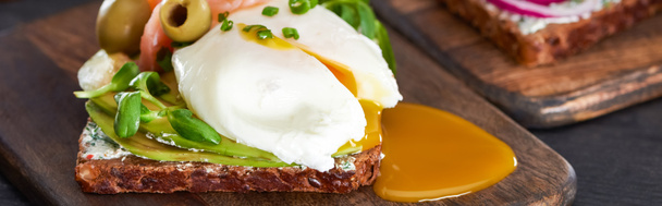 panoramic shot of smorrebrod sandwich with poached egg on wooden cutting board  - Photo, Image