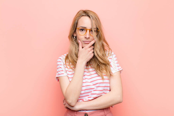 young pretty blonde woman looking serious, thoughtful and distrustful, with one arm crossed and hand on chin, weighting options against flat color wall - Foto, afbeelding