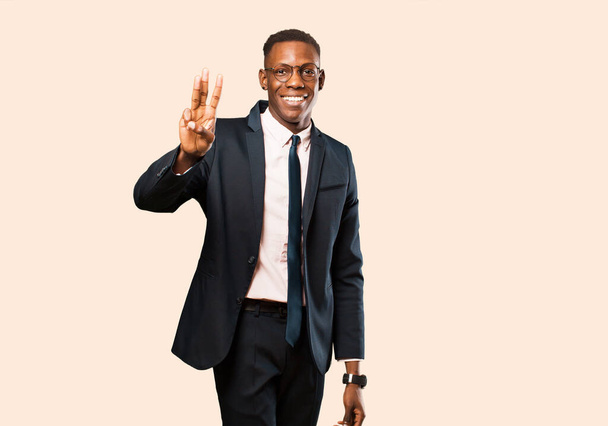 african american businessman smiling and looking friendly, showing number three or third with hand forward, counting down against beige wall - Foto, Bild