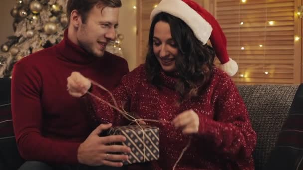Lovely Young Couple Finalizing together Christmas Gift Bandaging Tape and Tied in a Bow Sitting on the Sofa near Christmas Tree at Home Background. Concept of Holidays and New Year. - Záběry, video