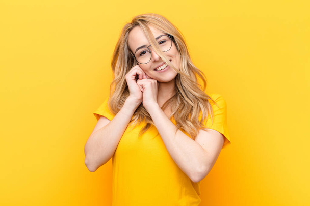 young pretty blonde woman feeling in love and looking cute, adorable and happy, smiling romantically with hands next to face against flat color wall - Foto, afbeelding