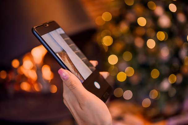 женская рука с маникюром с смартфоном и фотографией огня в камине на фоне рождественской елки в светильниках
 - Фото, изображение