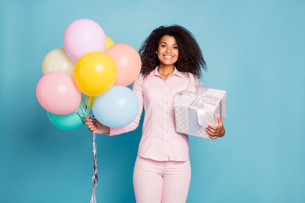 Photo of amazing wavy dark skin lady holding large wrapped giftbox and many colorful air balloons came to party wear pink shirt pants isolated blue color background - Zdjęcie, obraz