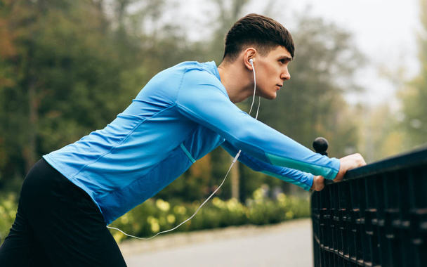 Side view image of young sportsman stretching outdoor in the morning on the fog nature background. Fitness male exercising in the park and listenting the music on earphones. People and sport concept - Foto, afbeelding