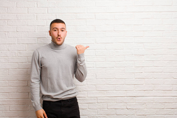young handsome man looking astonished in disbelief, pointing at object on the side and saying wow, unbelievable against flat background - Photo, Image