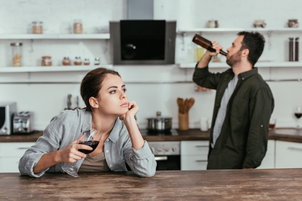 Selective focus of pensive woman with wine glass and husband drinking beer at kitchen - Fotoğraf, Görsel