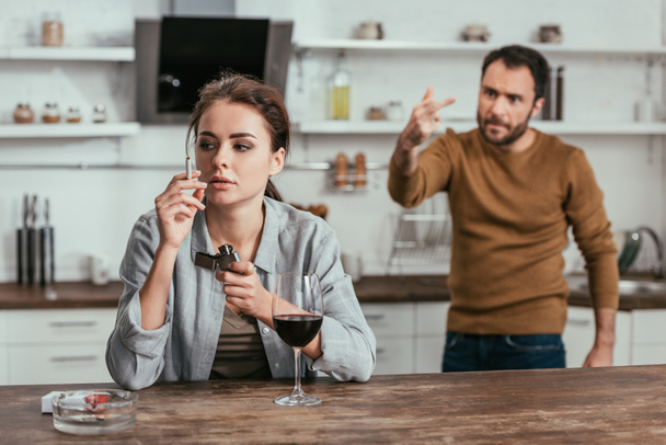 Selective focus of woman with wine smoking while angry husband showing middle finger - Zdjęcie, obraz