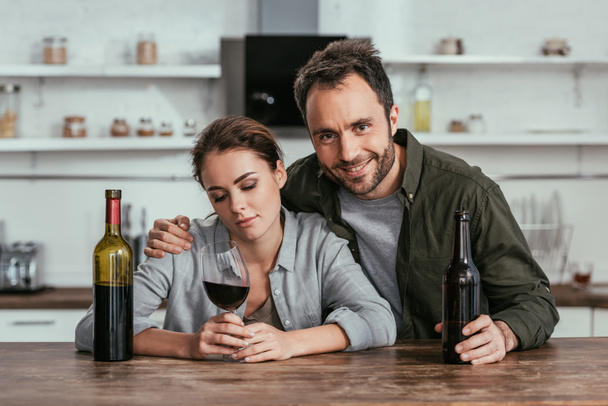 Улыбающийся мужчина с пивом обнимает грустную женщину с вином на кухне
 - Фото, изображение