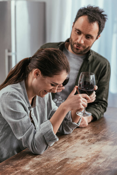 Enfoque selectivo de pareja con copas de vino riendo en la cocina
 - Foto, Imagen
