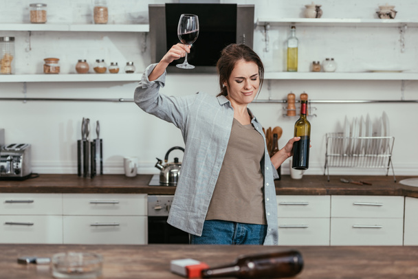 Enfoque selectivo de la mujer borracha sosteniendo vino con botella de cerveza y cigarrillos en la mesa de la cocina
 - Foto, Imagen