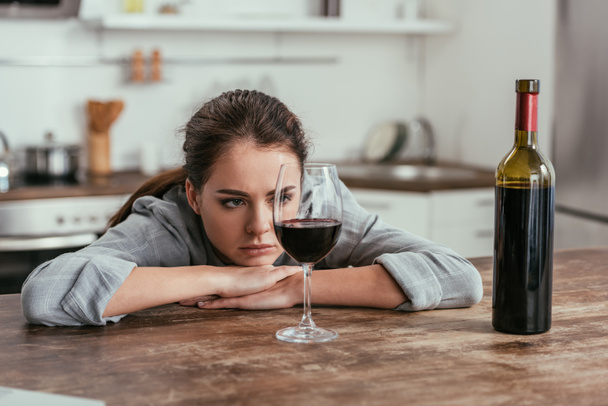 Thoughtful woman looking at wine glass on kitchen table - Foto, imagen