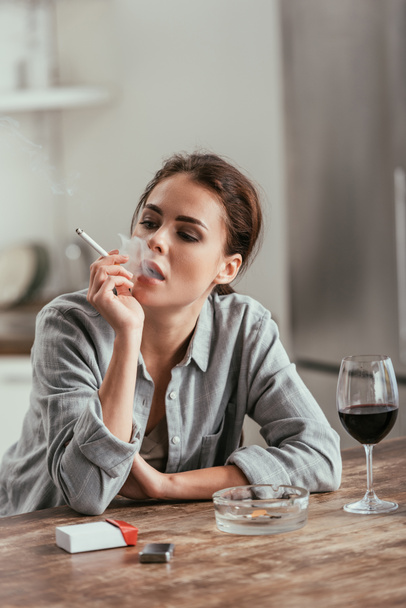 Женщина курит сигарету рядом с бокалом вина на столе
 - Фото, изображение