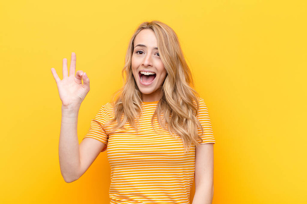 young pretty blonde woman feeling successful and satisfied, smiling with mouth wide open, making okay sign with hand against flat color wall - 写真・画像