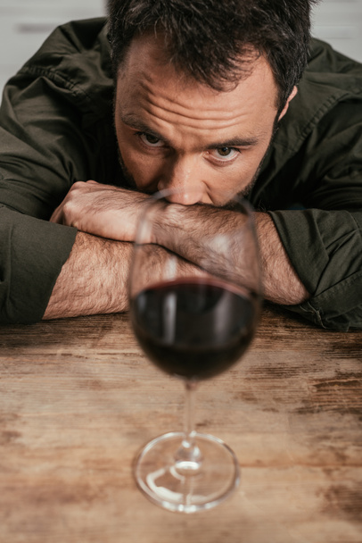 Selektywne skupienie się na uzależnionym od alkoholu człowieku patrzącym na kieliszek wina na stole - Zdjęcie, obraz