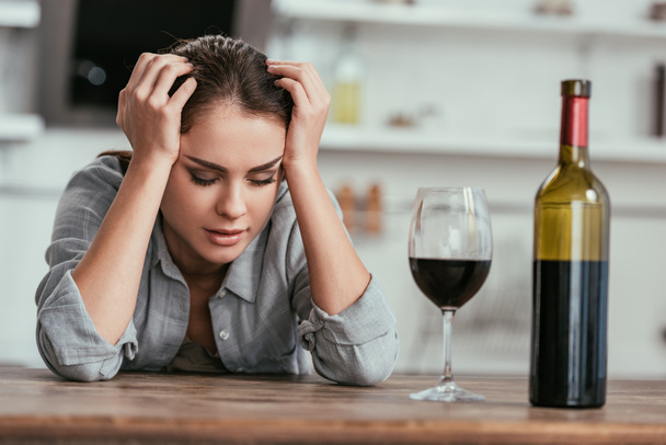 Selective focus of woman with alcohol addiction sitting beside wine on kitchen table - Foto, imagen