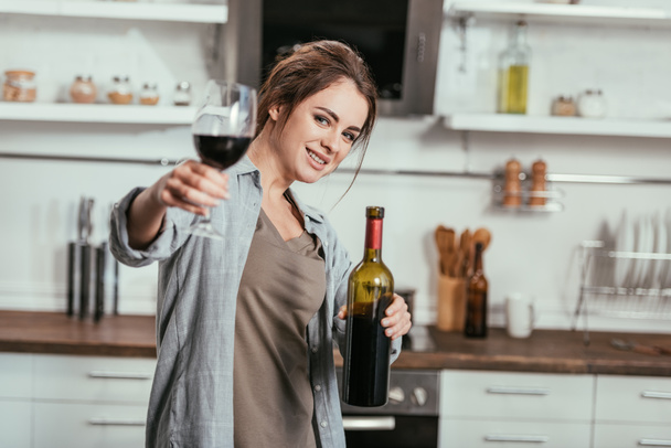 Selective focus of smiling woman holding bottle and wine glass at kitchen - Foto, Bild