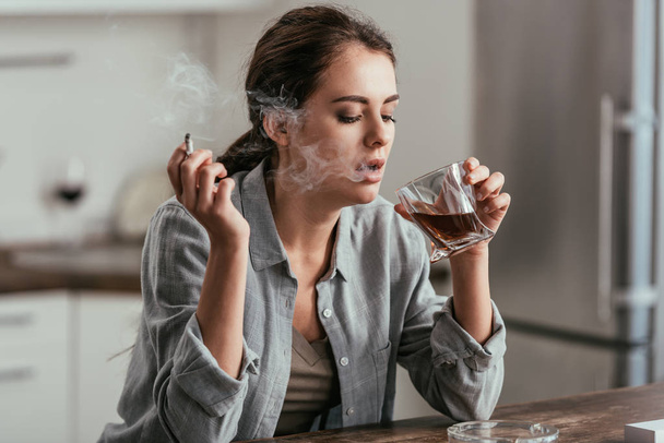 Vrouw roken en kijken naar whiskey glas aan keukentafel - Foto, afbeelding