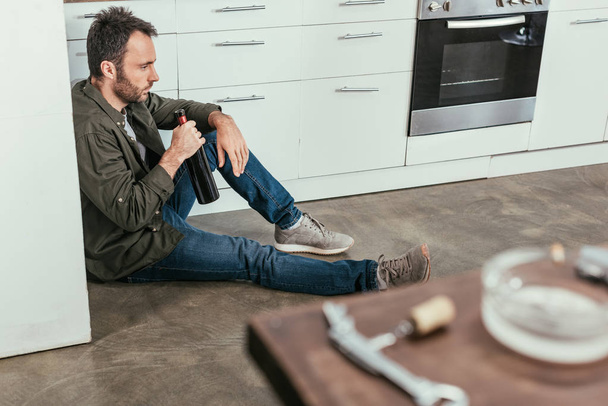Selective focus of sad man with wine bottle sitting on kitchen floor - Photo, Image