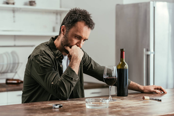 Pensive man looking at wine glass and bottle on kitchen table - Valokuva, kuva