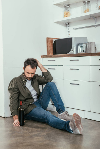 Hombre molesto sentado en el suelo de la cocina con botella de vino
 - Foto, Imagen