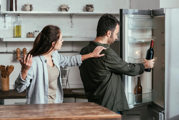 Angry woman quarreling with husband taking wine bottle from fridge - 写真・画像