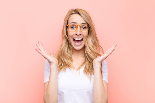 young pretty blonde woman looking happy and excited, shocked with an unexpected surprise with both hands open next to face against flat color wall - Photo, Image