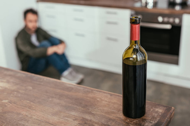 Enfoque selectivo de la botella de vino en la mesa y el hombre sentado en el suelo de la cocina
 - Foto, Imagen