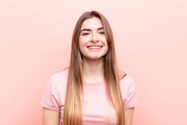 young pretty woman looking happy and goofy with a broad, fun, loony smile and eyes wide open against pink wall - Foto, afbeelding