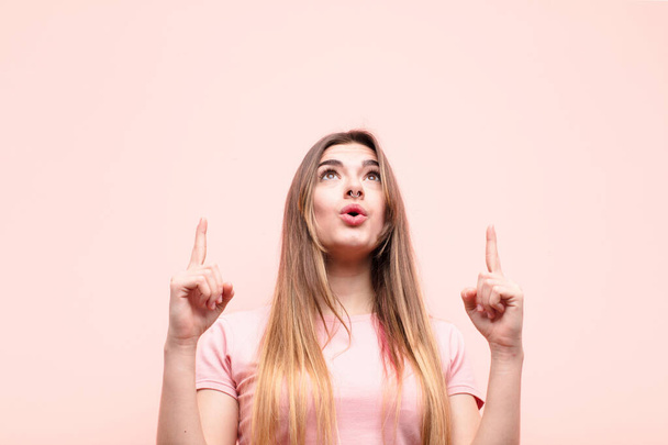 young pretty woman looking shocked, amazed and open mouthed, pointing upwards with both hands to copy space against pink wall - Foto, immagini