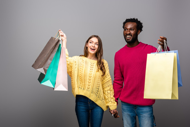 happy interracial couple in sweaters holding hands and shopping bags on grey background - Fotó, kép
