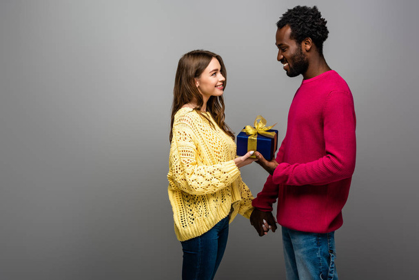 happy interracial couple in sweaters holding present on grey background - Фото, изображение