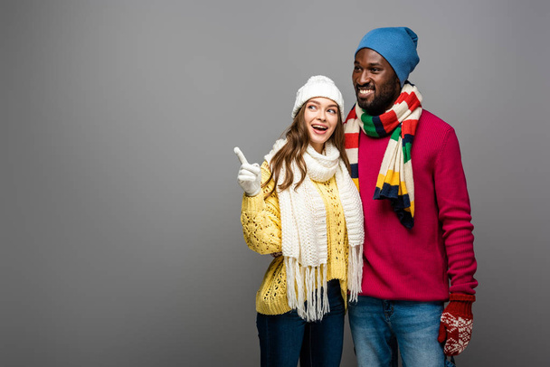 smiling interracial couple in winter outfit hugging and pointing with finger on grey background - Foto, immagini