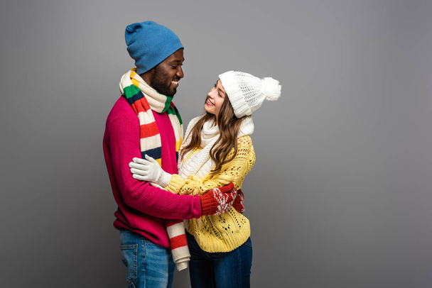 glimlachen interraciale paar in de winter outfit knuffelen op grijze achtergrond - Foto, afbeelding