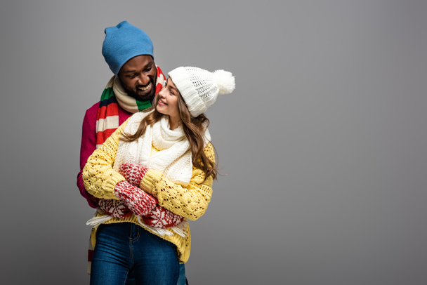 happy interracial couple in winter outfit hugging on grey background - Foto, imagen