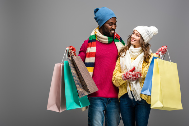 happy interracial couple in winter outfit holding shopping bags on grey background - Foto, immagini