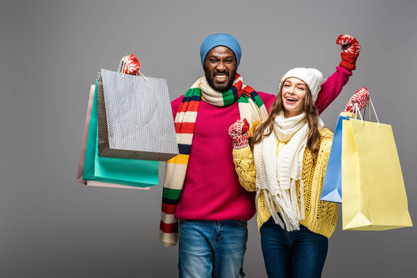 happy interracial couple in winter outfit holding shopping bags on grey background - Фото, зображення